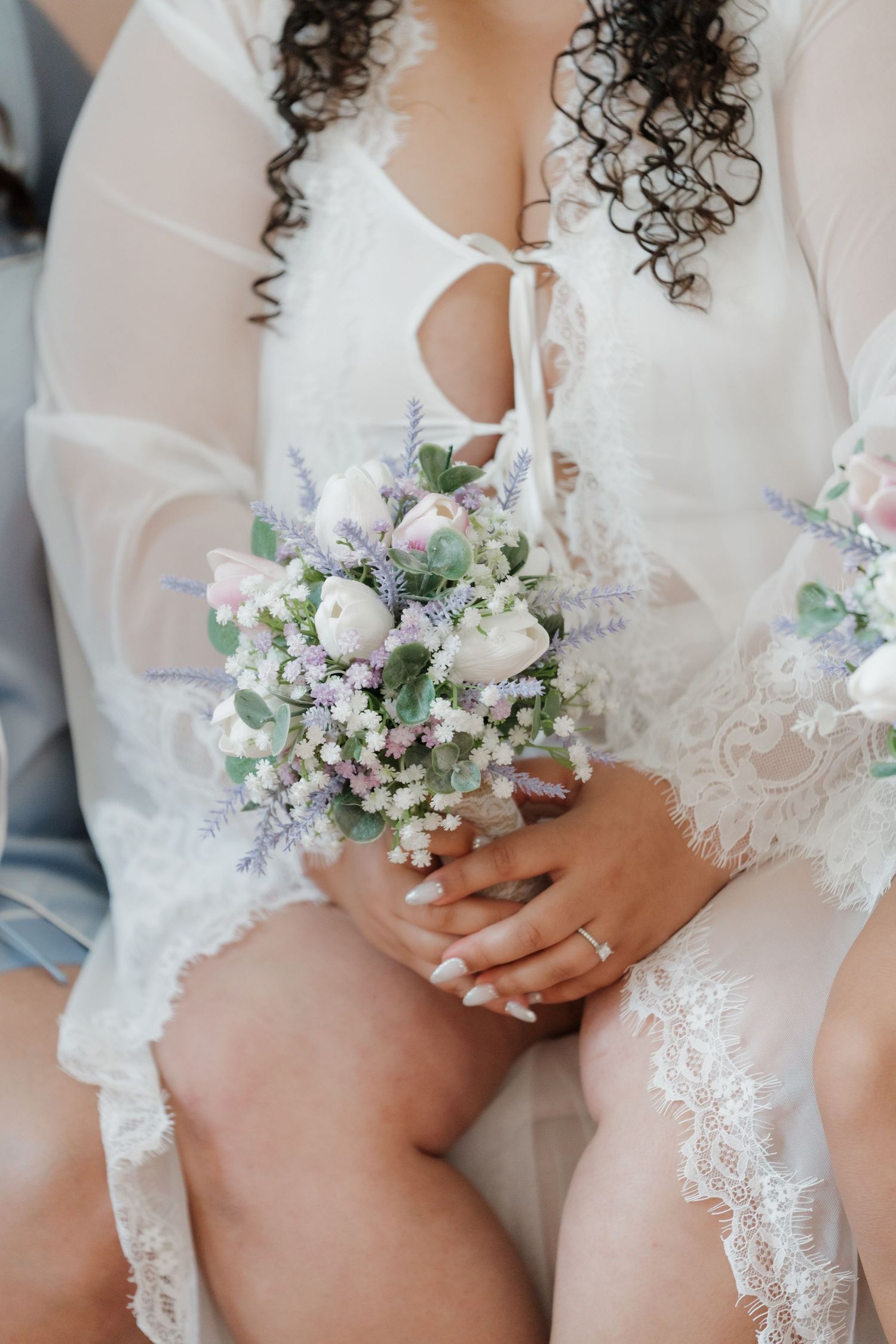 Bride in white lace robe holding small bouquet of white roses, lavender, and gypsophila flowers.