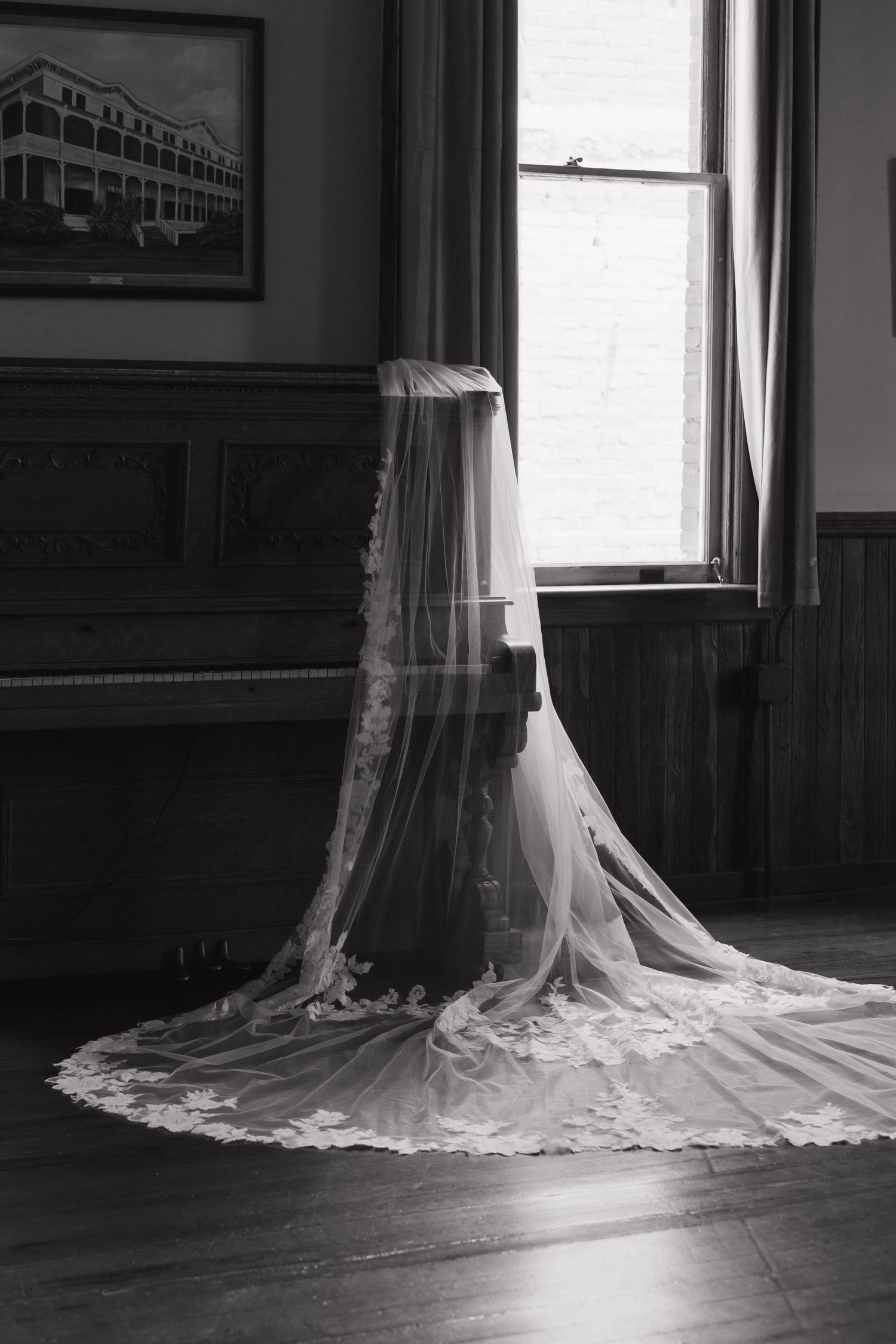 Black and white photo of a lace wedding veil draped over a stand near a piano and window in a vintage room.