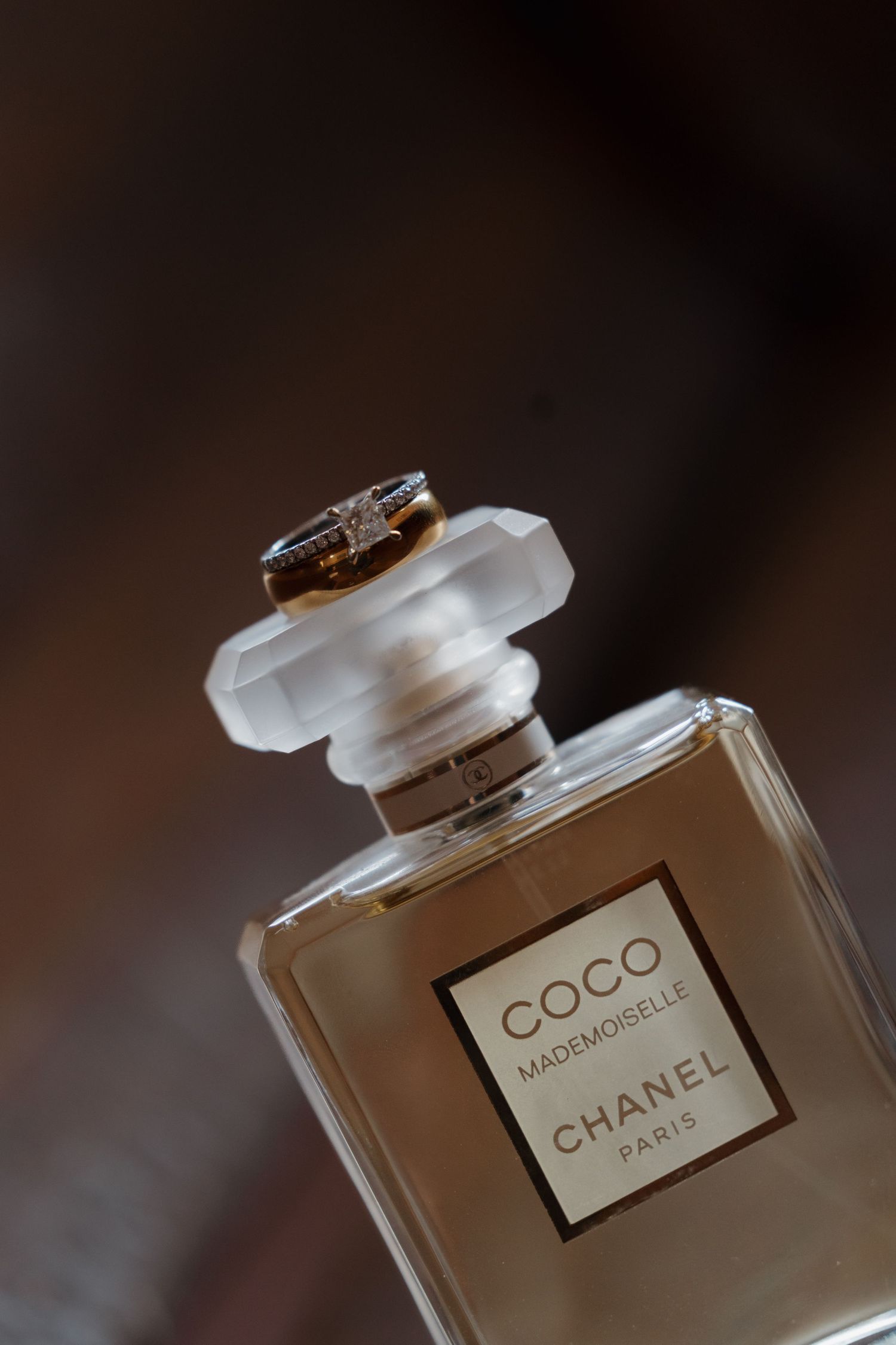 Wedding rings rest atop Coco Mademoiselle Chanel Paris perfume bottle against dark background.