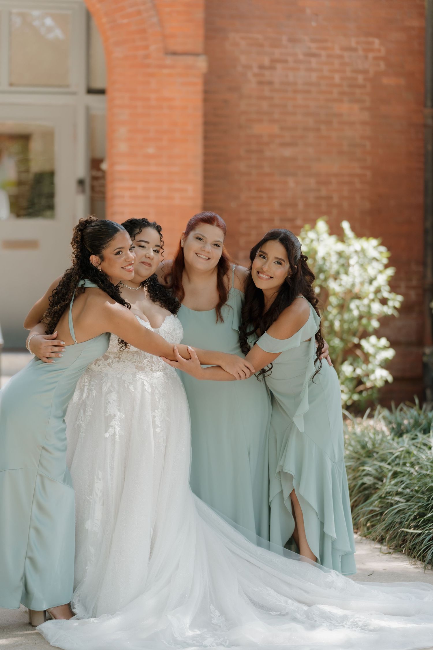 Bride in white lace gown poses with three bridesmaids in sage green dresses outside brick building.
