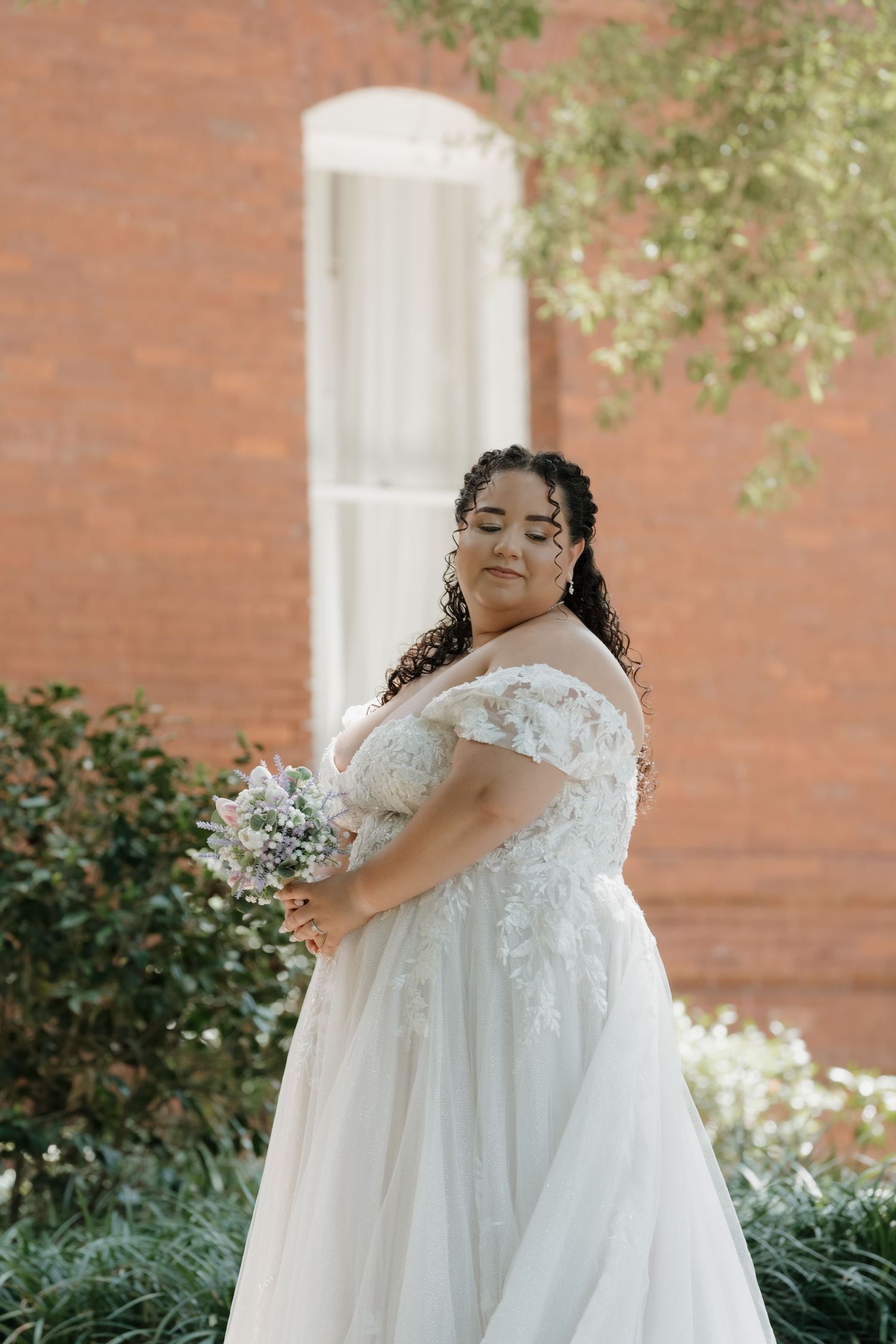 Bride in off-shoulder lace wedding gown holding bouquet outdoors near brick building.