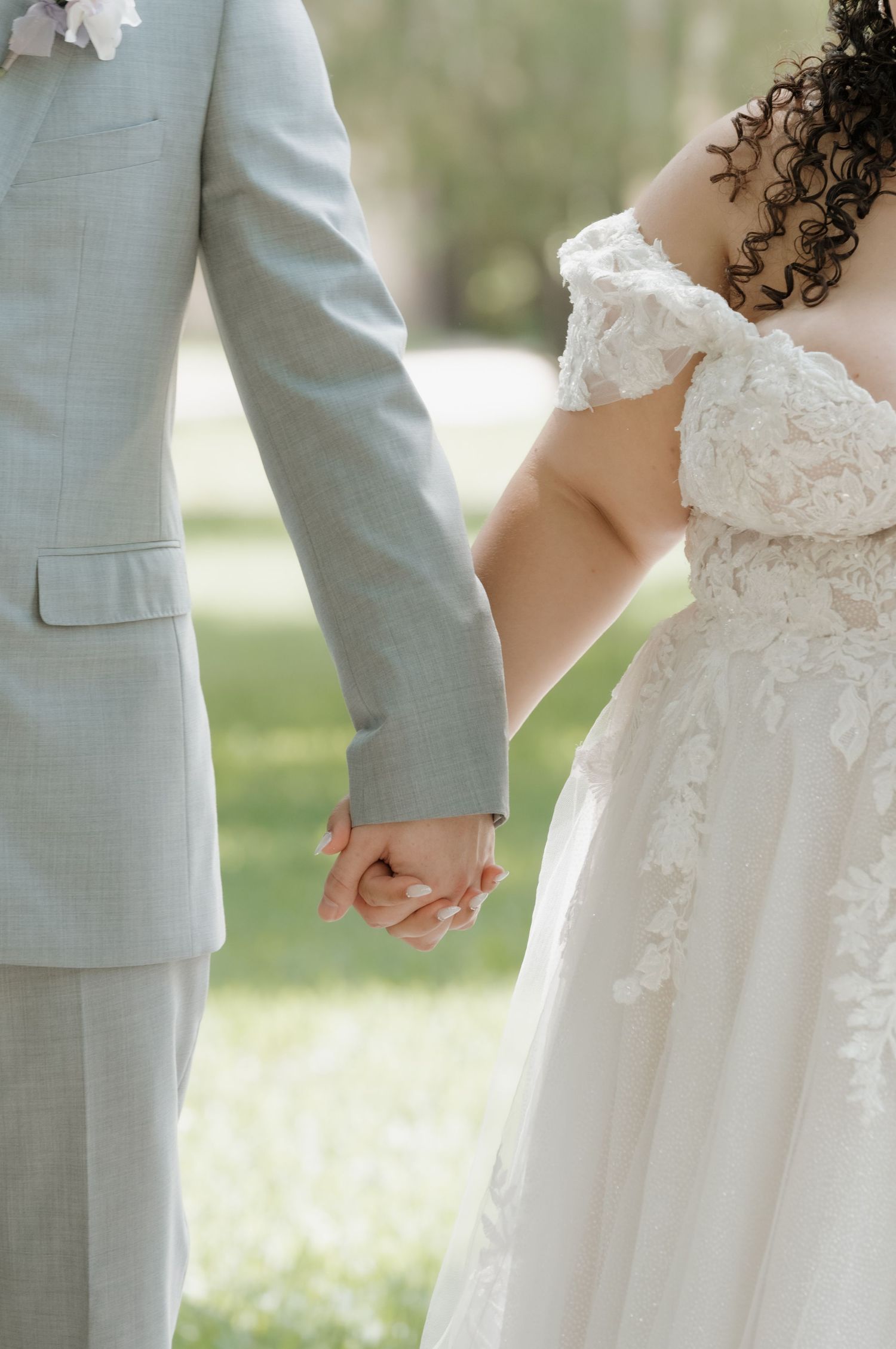 Bride and groom holding hands outdoors, bride in lace off-shoulder gown, groom in light gray suit.