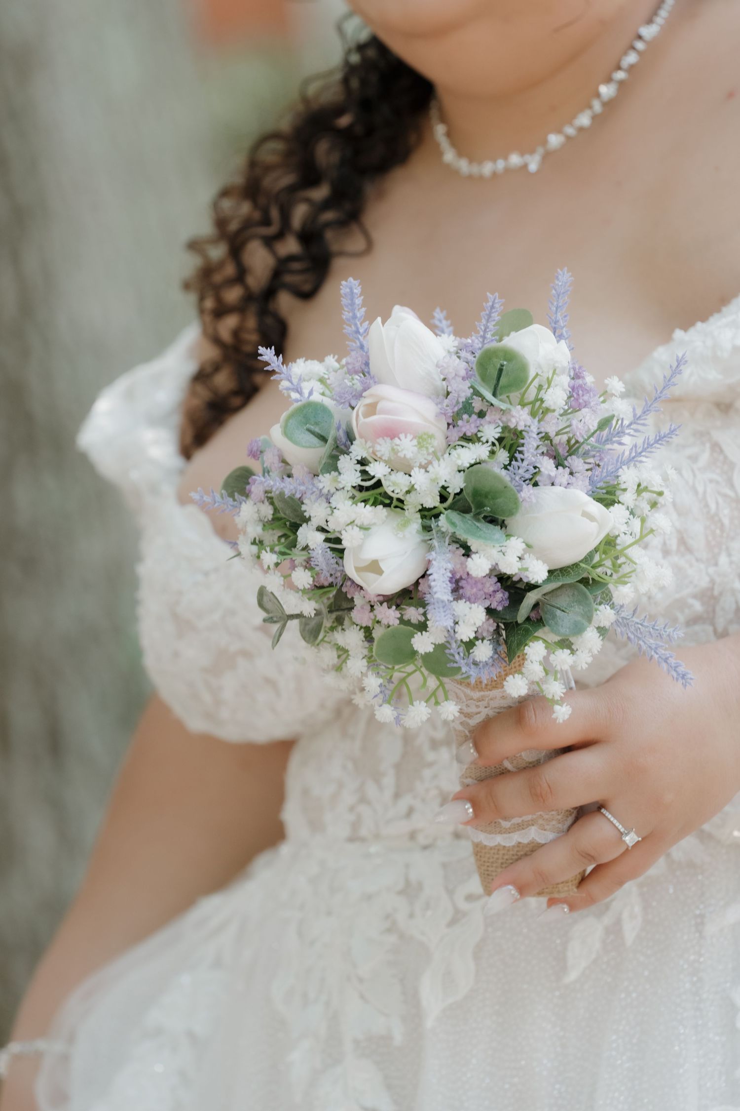 Bride in lace wedding dress holding a bouquet of white tulips, lavender, and eucalyptus, wearing a pearl necklace.