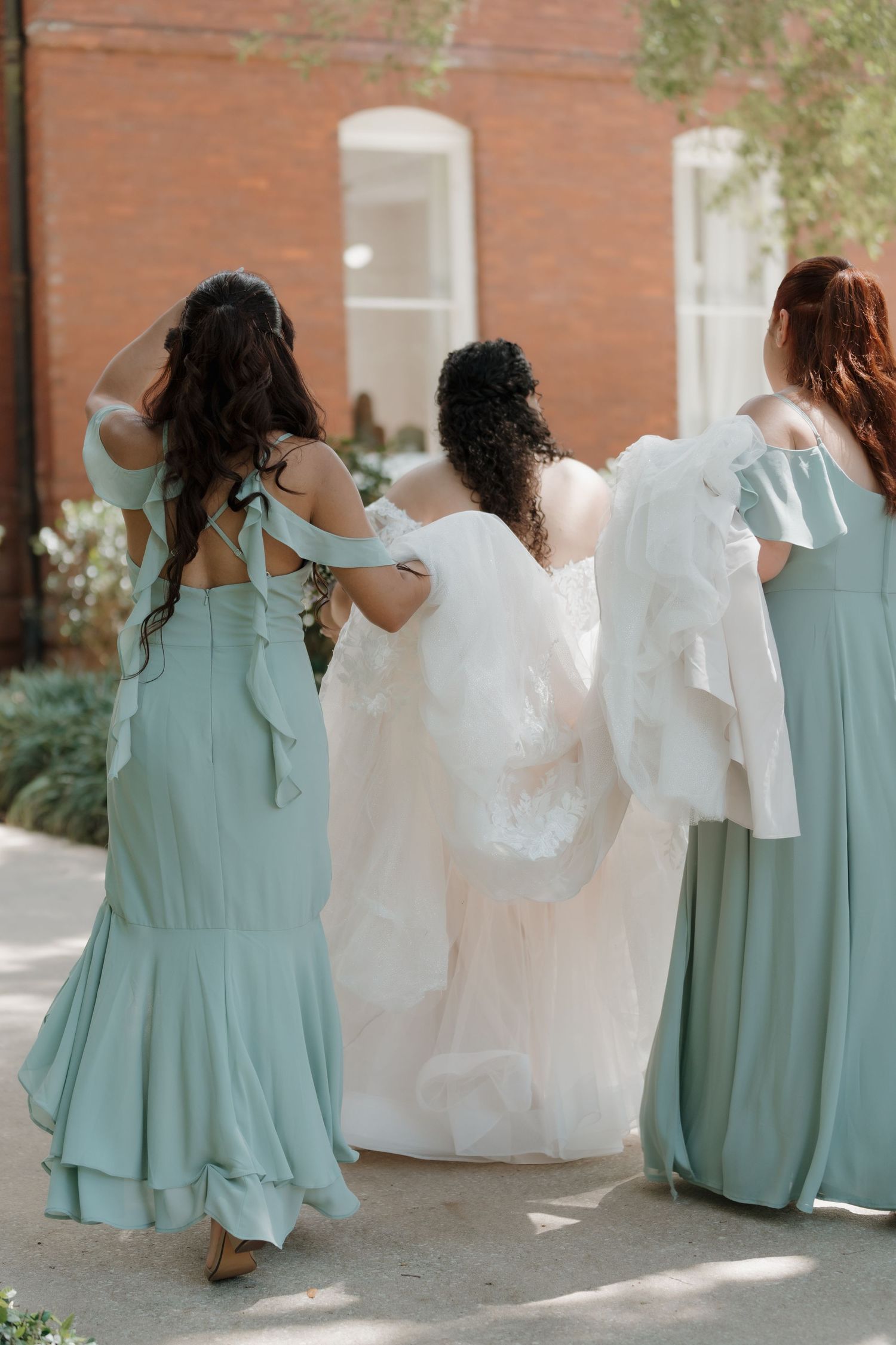 Bridesmaids in sage green dresses helping bride with white wedding gown train while walking outdoors.