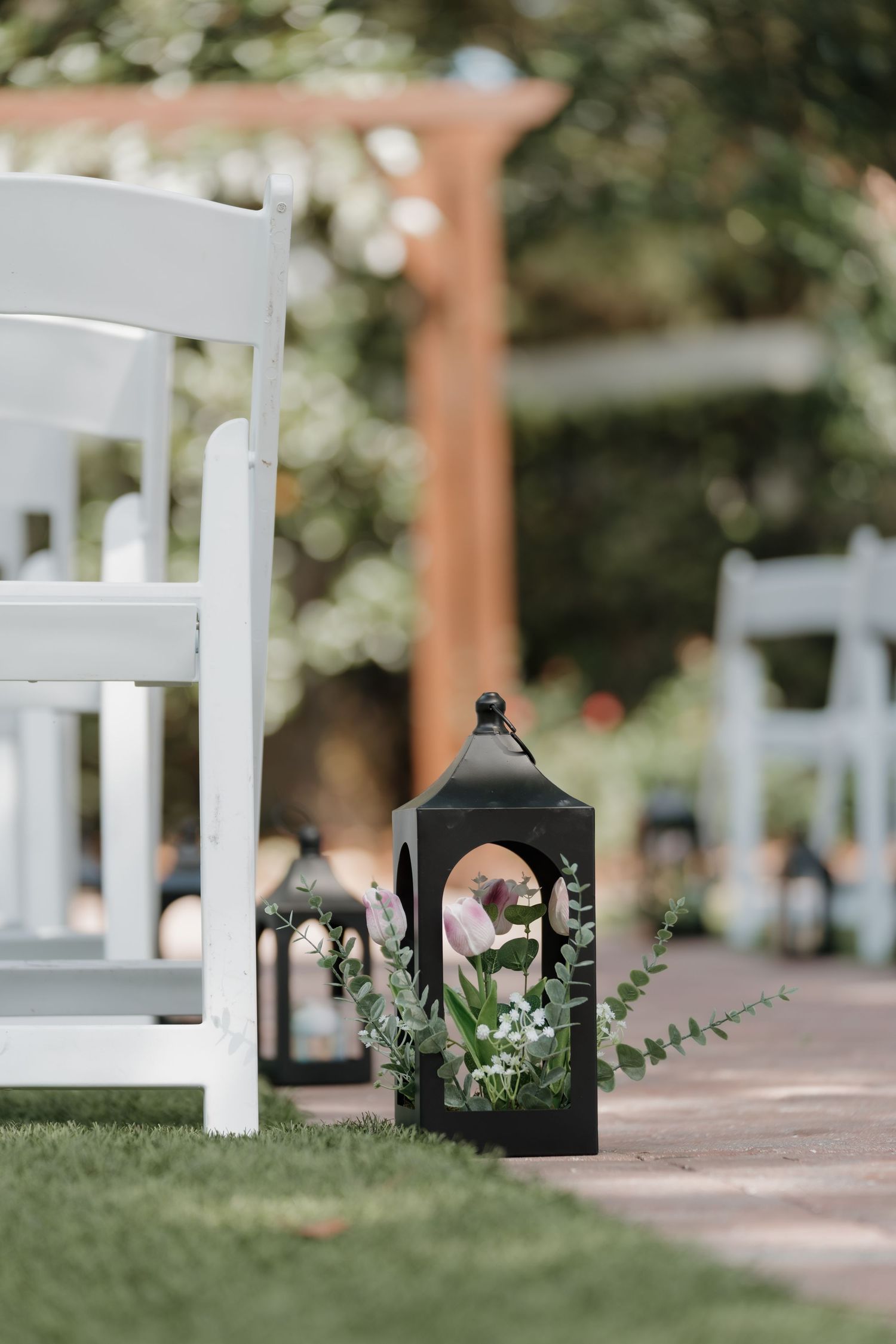 Black lantern with pink flowers and eucalyptus decorates outdoor wedding ceremony aisle beside white chairs.
