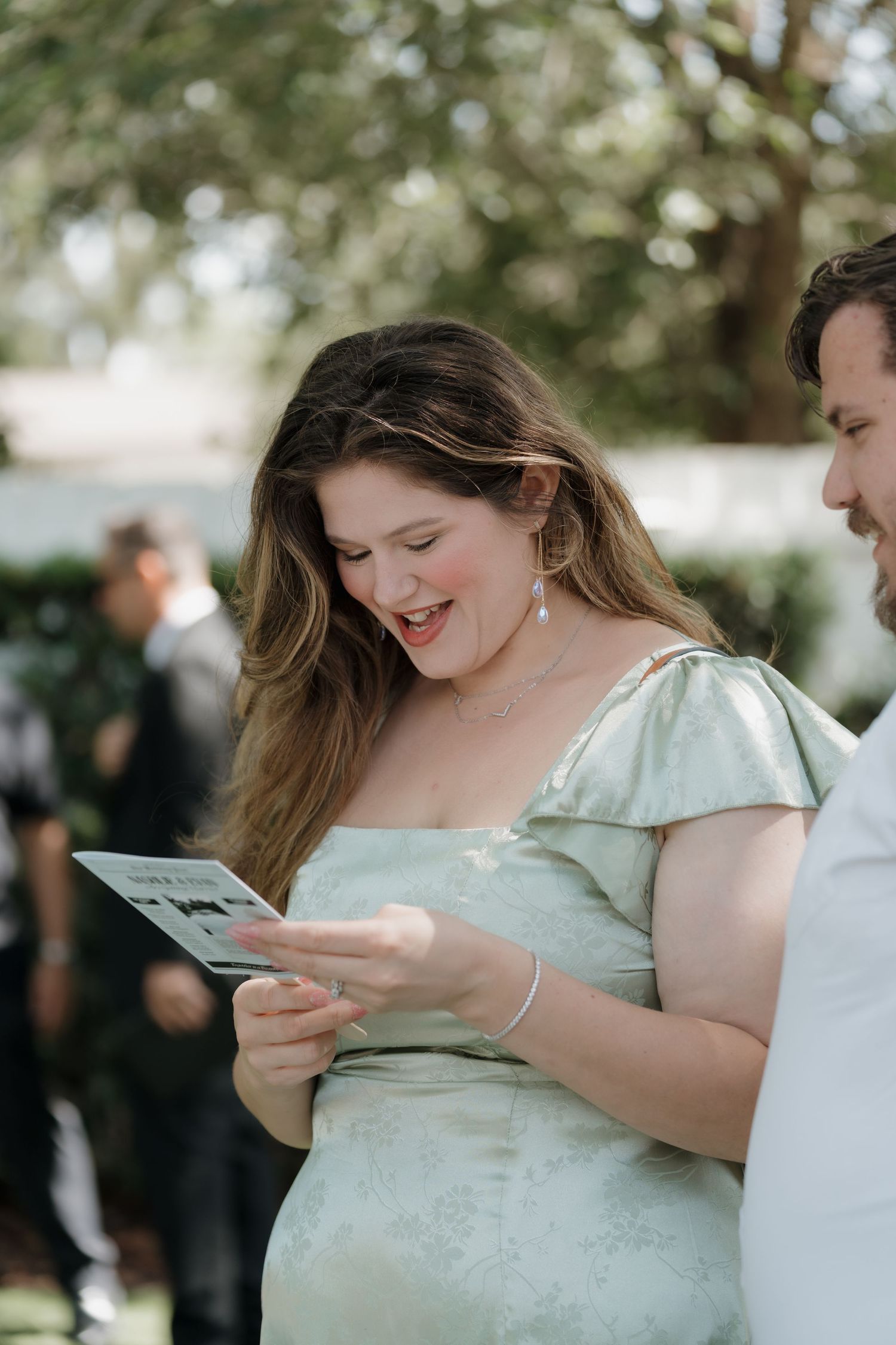 Pregnant wedding guest in sage green dress smiling while reading ceremony program outdoors.