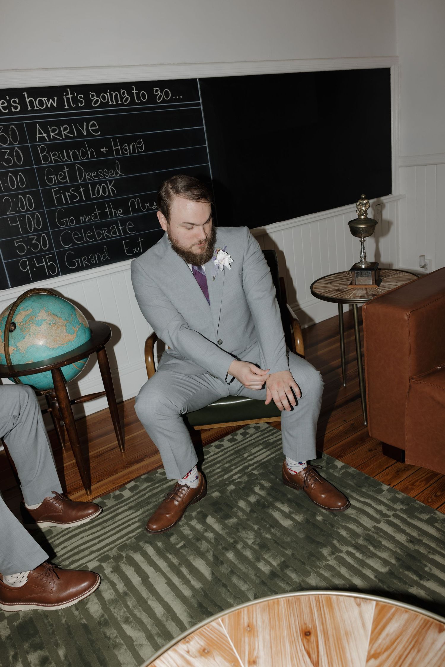 Groom in gray suit sitting pensively in a vintage room with globe and chalkboard timeline.