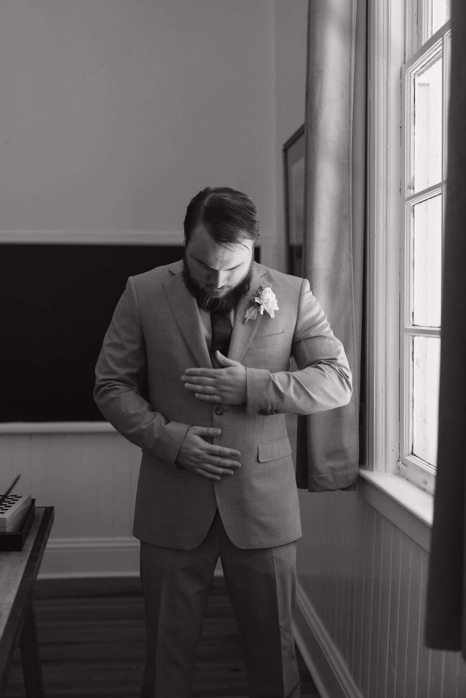 Groom in grey suit adjusting jacket near window in black and white wedding preparation photo.