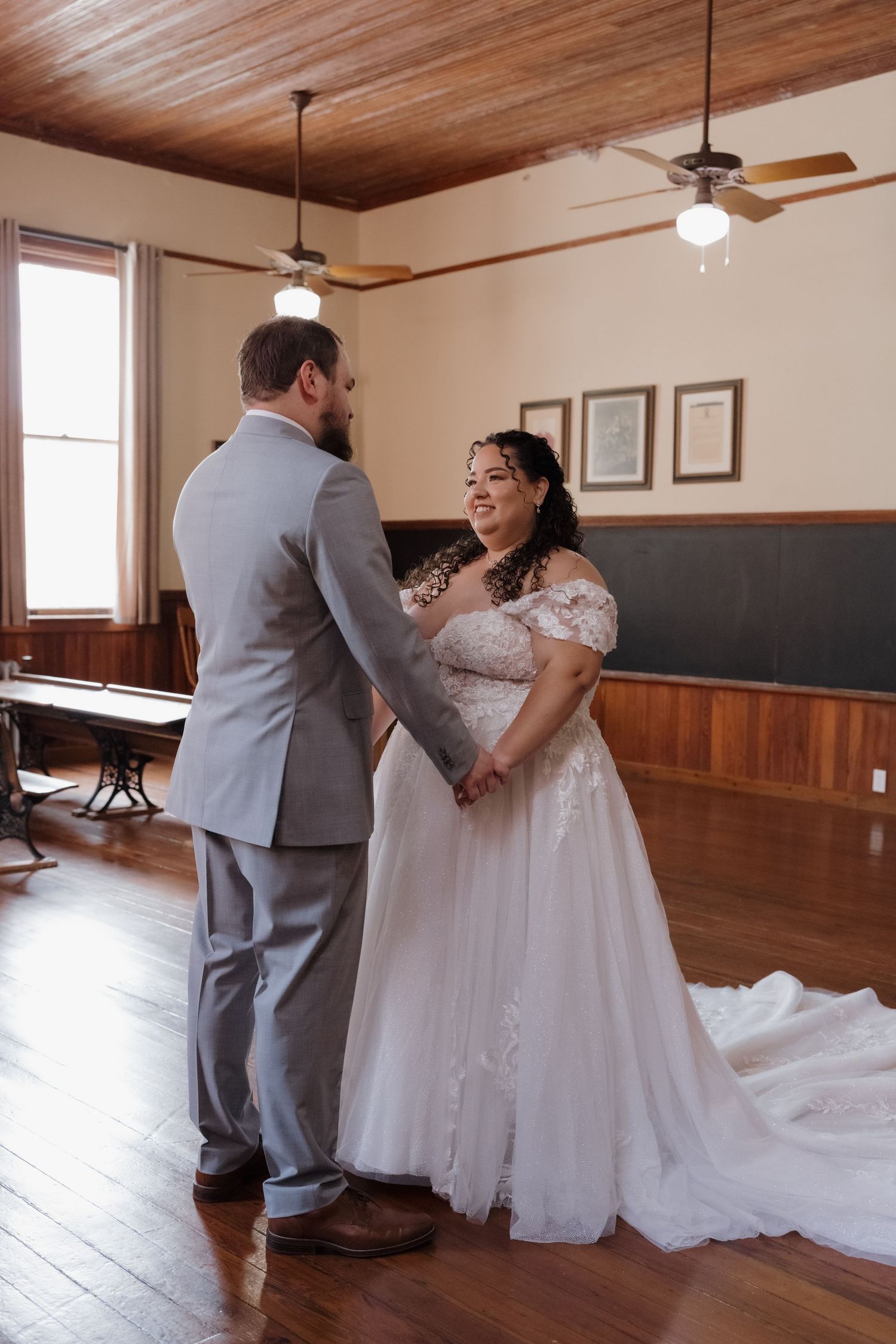 Bride in lace ball gown holds hands with groom in gray suit during first look in rustic venue.