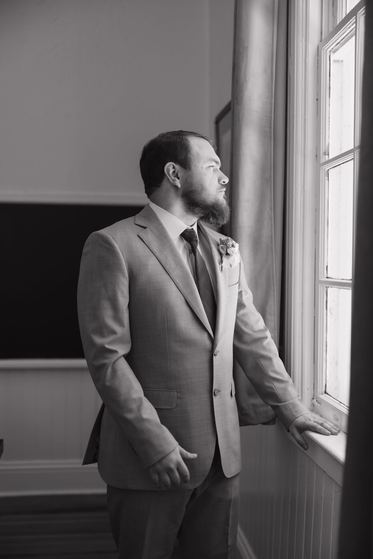 Groom in gray suit with boutonniere gazing out a window in black and white wedding photo.