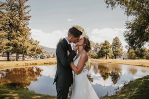 A romantic couple embraces by a serene lake during sunset, surrounded by lush trees and golden sunlight.