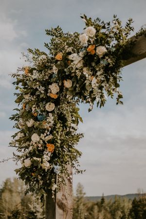 A rustic wooden arch decorated with lush white roses and greenery against a cloudy sky for an outdoor wedding ceremony.
