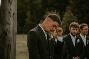 Groomsmen in black suits and ties share an emotional moment during an outdoor wedding ceremony.