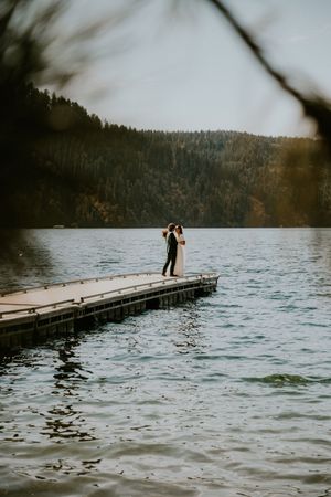 A couple stands together on a wooden dock overlooking a serene lake surrounded by forested hills on a moody day.