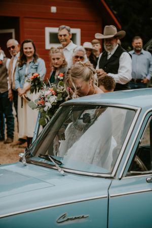 A vintage blue classic car decorated with flowers serves as a backdrop at a rustic outdoor wedding celebration.