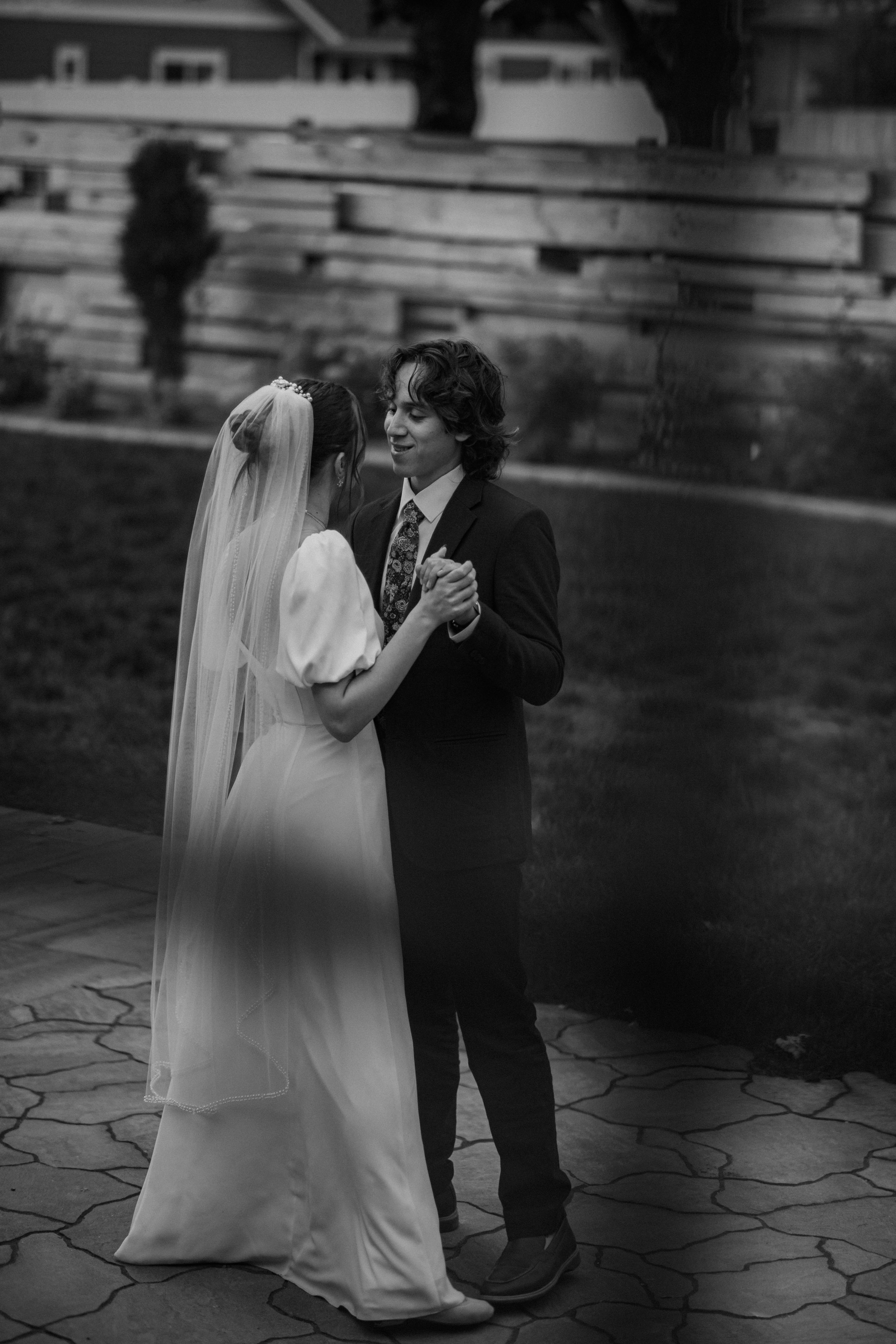 A black and white wedding photograph showing a couple sharing their first dance on a stone patio.