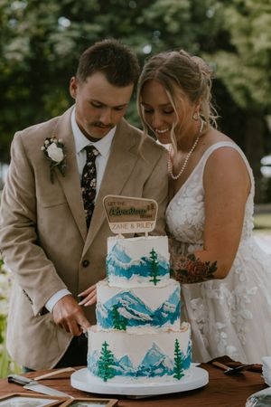 Newlyweds cut their elegant three-tier wedding cake decorated with turquoise mountain landscape design.