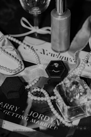 Black and white wedding details including jewelry, pearls, perfume bottle and wedding stationery arranged on a surface.