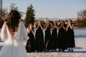 Bridesmaids in black dresses react with surprise to bride in white dress during winter photoshoot in snowy setting.