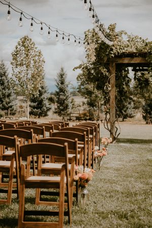 Outdoor wedding ceremony setup with wooden chairs and string lights under a wooden arch surrounded by trees.