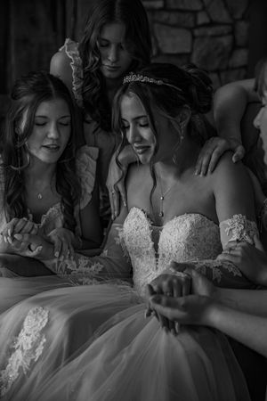 Black and white image shows bride and bridesmaids in elegant white lace dresses sitting together during a wedding event.