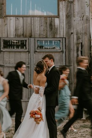 Wedding party in motion blur stands against rustic wooden barn wall during outdoor autumn celebration.