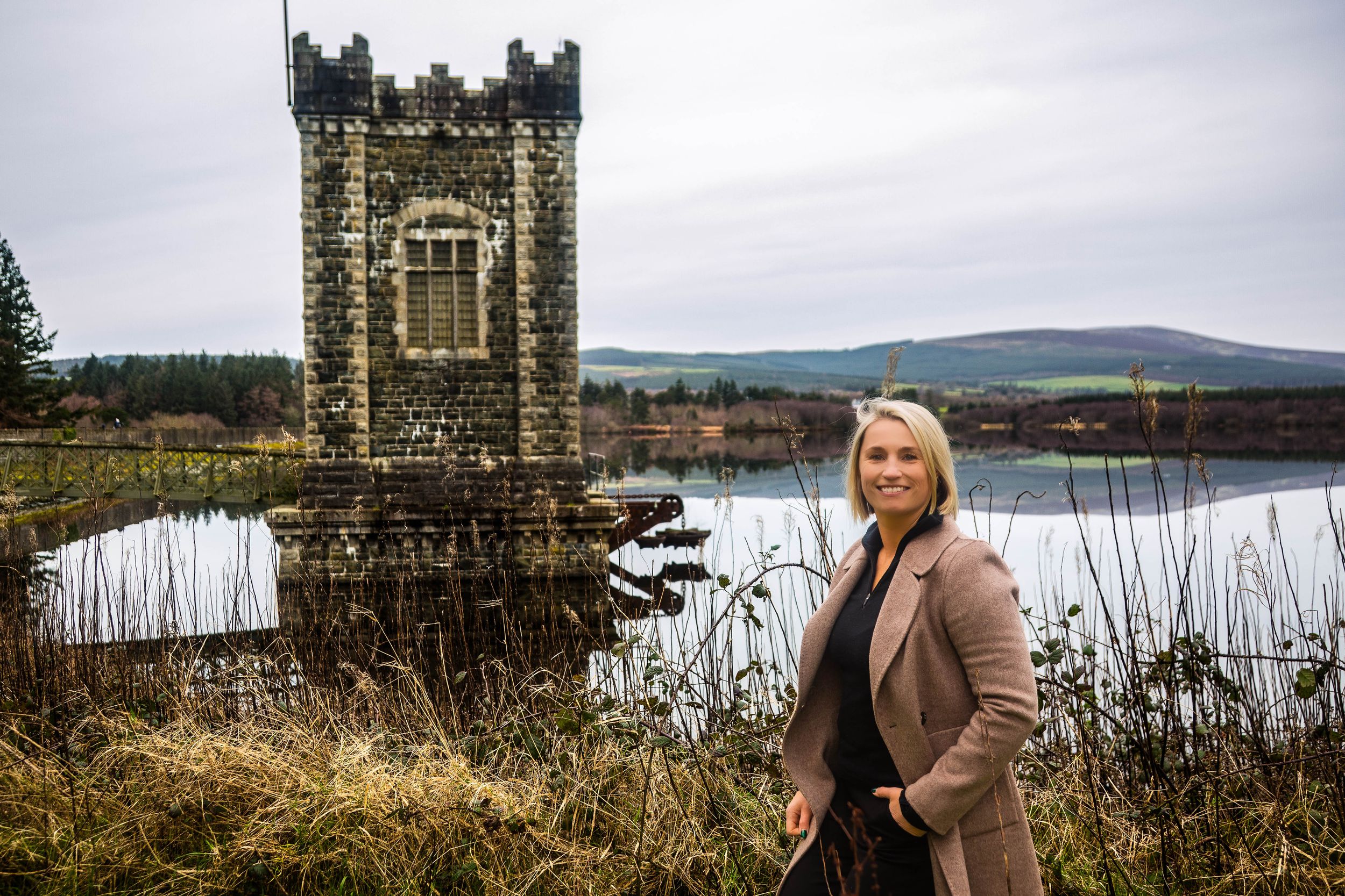 Vartry Reservoir Draw Tower stands majestically beside tranquil lake with Wicklow mountains in distance and lady posing in the front
