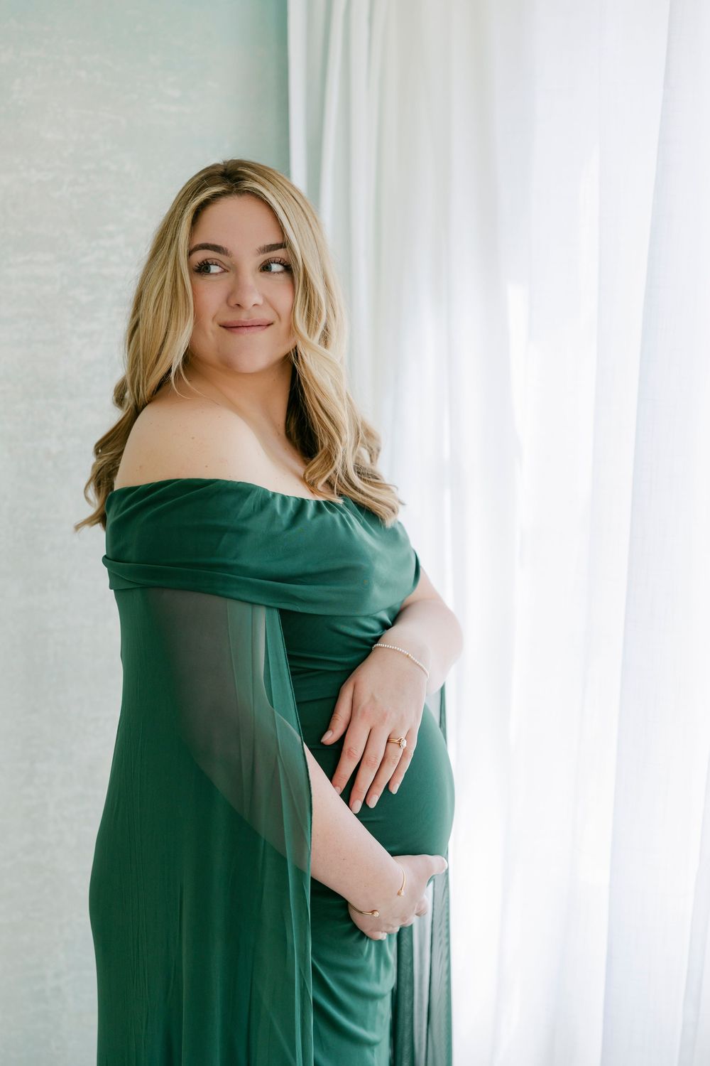 Woman in elegant emerald green off-shoulder dress poses against white curtain background in studio portraits.