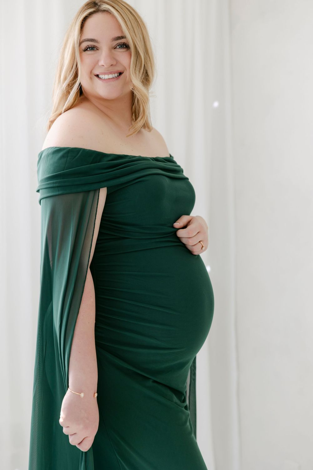 Maternity photo series showing a woman in an elegant emerald green off-shoulder dress against a white backdrop.