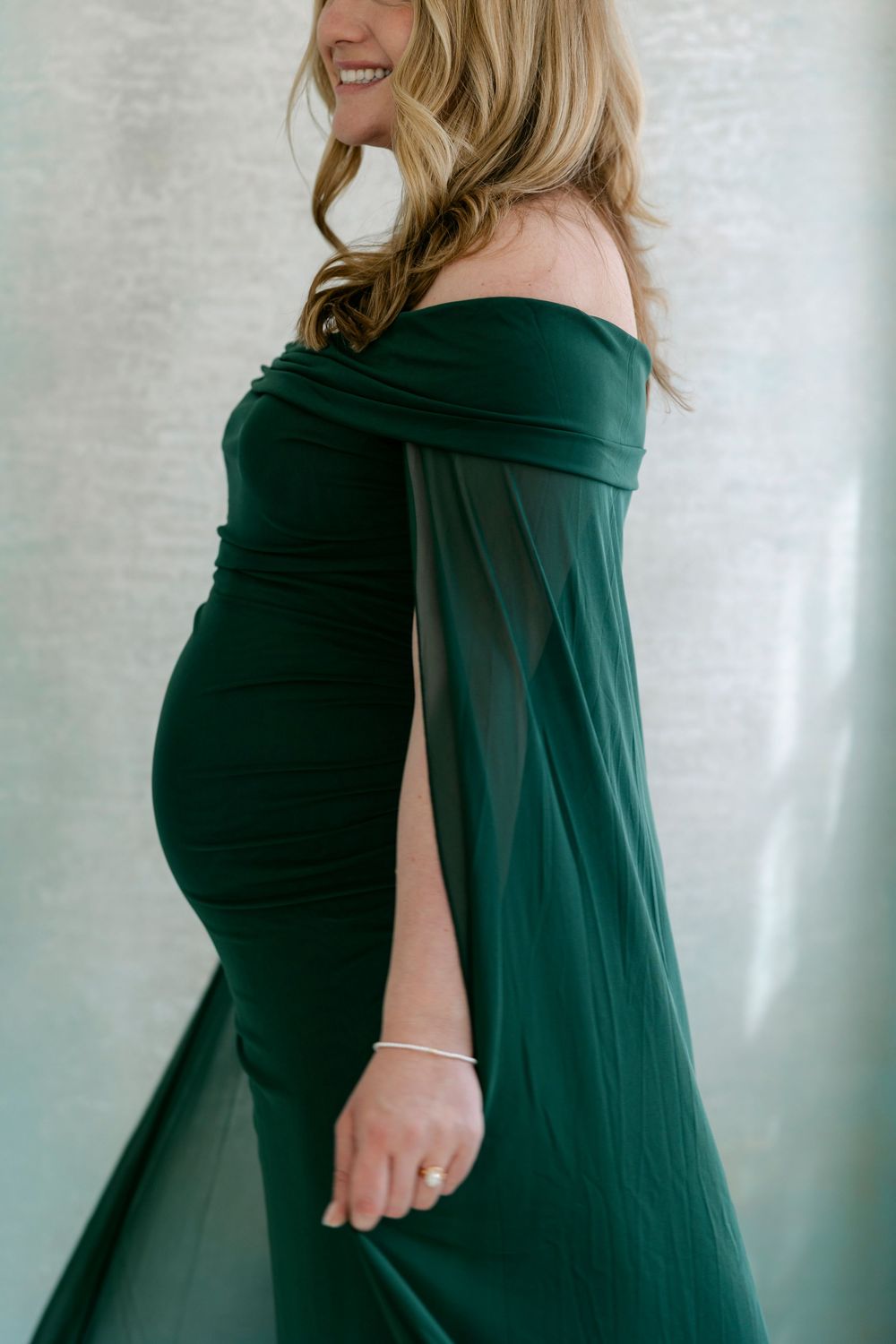 Elegant emerald green off-shoulder maternity gown with flowing fabric drapes gracefully over a baby bump.