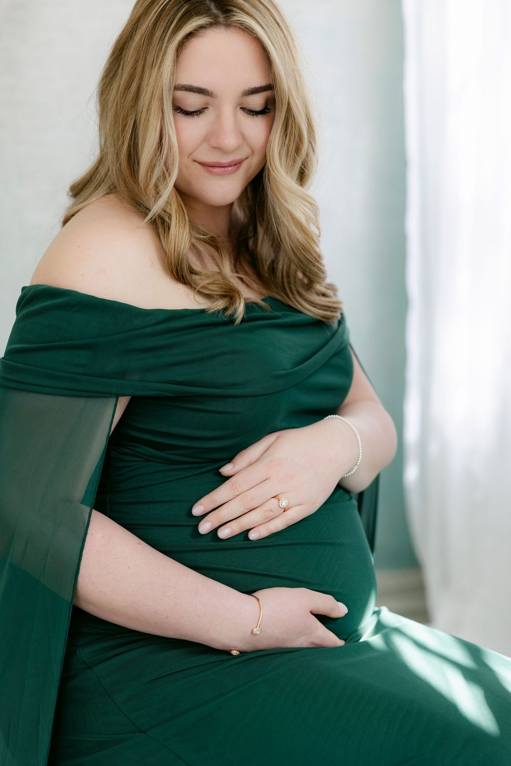 Pregnant woman in emerald green dress poses for maternity photos in natural window light.
