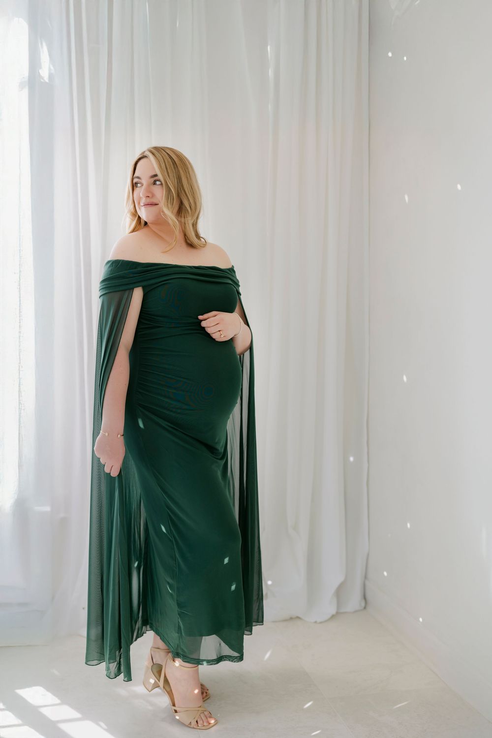 Elegant emerald green off-shoulder evening gown with flowing cape detail and nude heels in a bright studio setting.