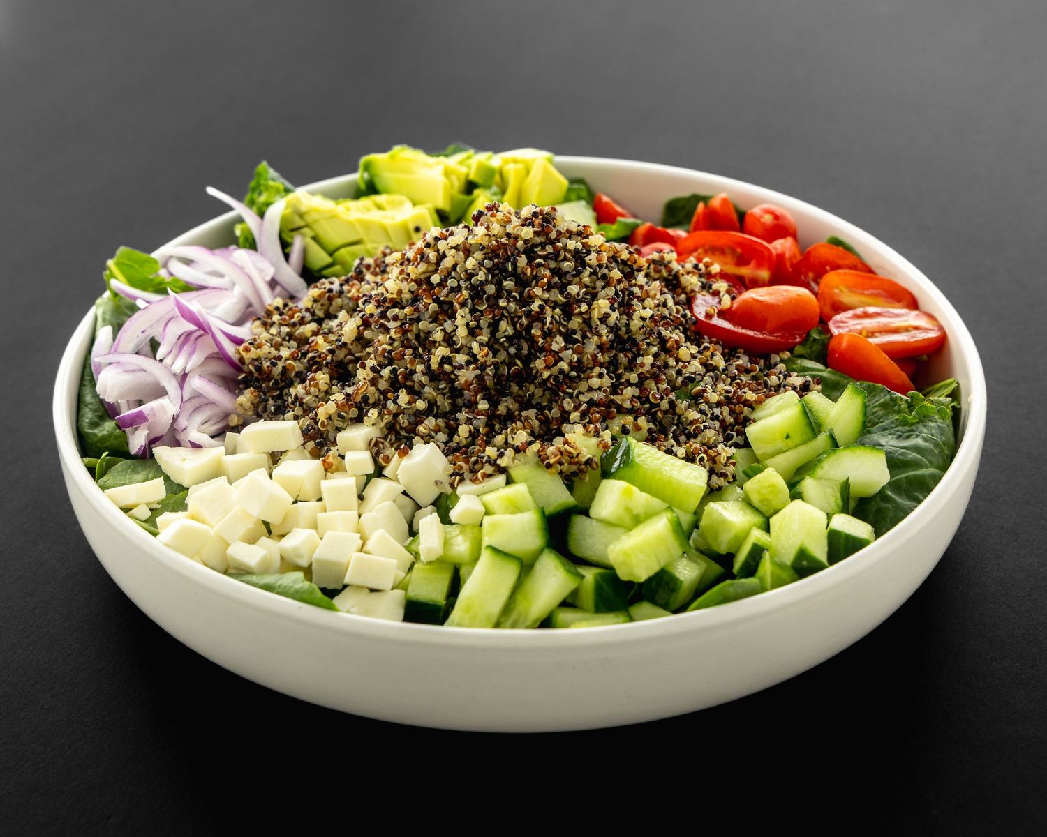 Fresh quinoa bowl with chopped vegetables including cucumber, tomatoes, red onion, and yellow peppers in white dish.