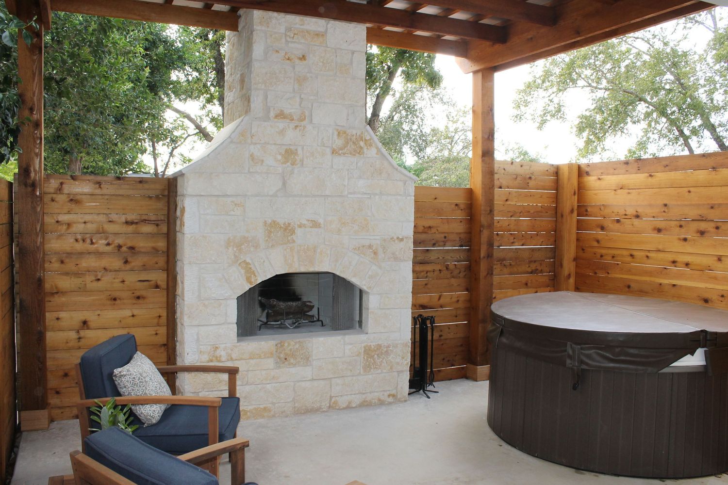 Outdoor living space with limestone fireplace and hot tub under a wooden pergola.