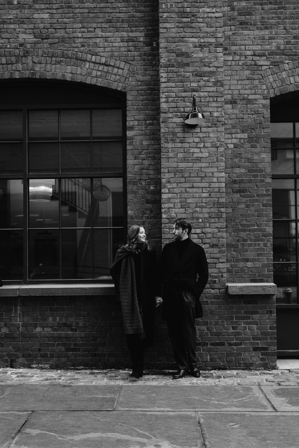 Two people in dark clothing stand against a brick wall in a black and white urban scene.
