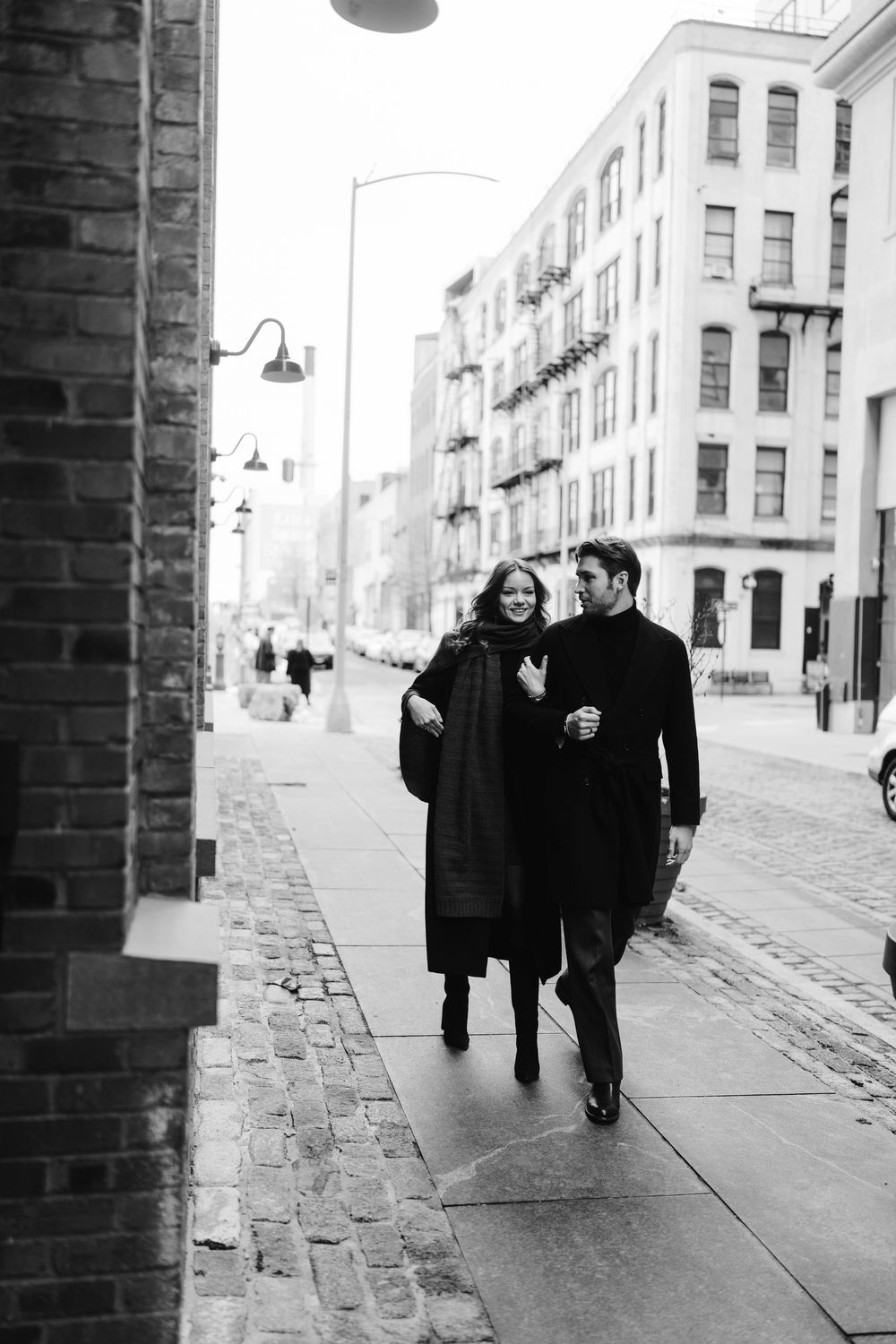 Two people walking together on a cobblestone street in a black and white vintage urban scene.