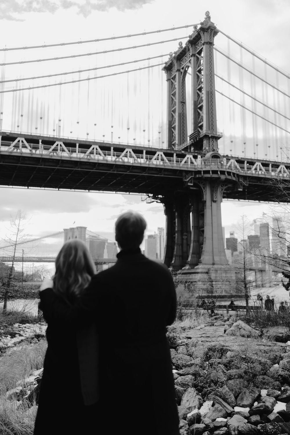 A silhouetted couple admires the Manhattan Bridge in a moody black and white photograph of New York City.