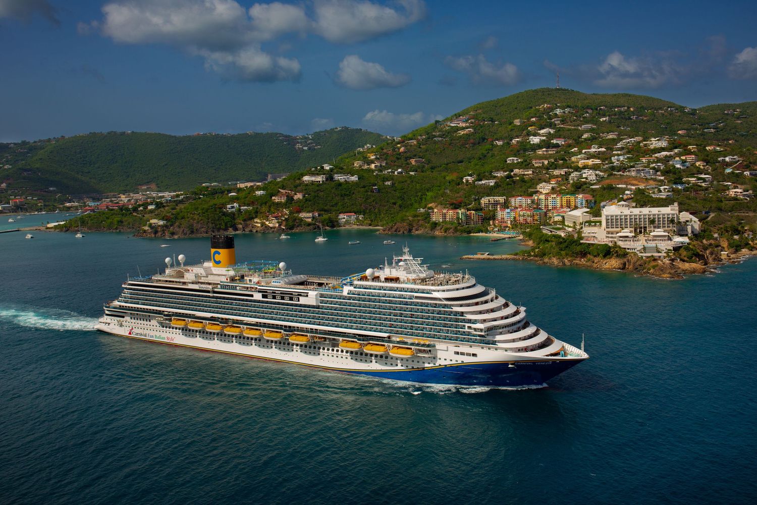 Large cruise ship with blue and white exterior sailing near a tropical hillside town with turquoise Caribbean waters.