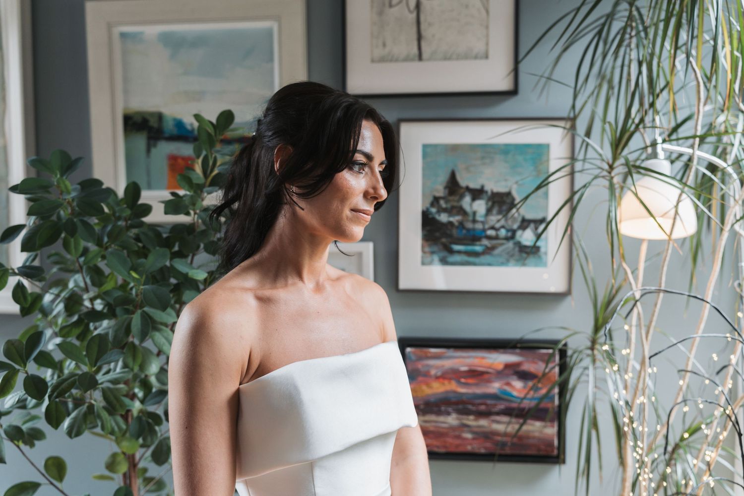 A person in a white strapless dress stands in front of framed artwork and houseplants in a bright interior space.