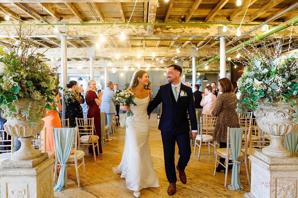 A couple walks down the aisle in a rustic venue decorated with greenery and white chairs.