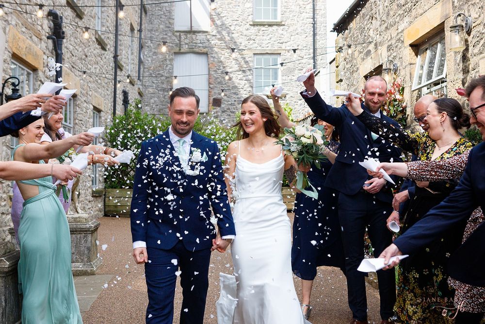 A joyful wedding couple walks through confetti being thrown by guests outside a stone building.