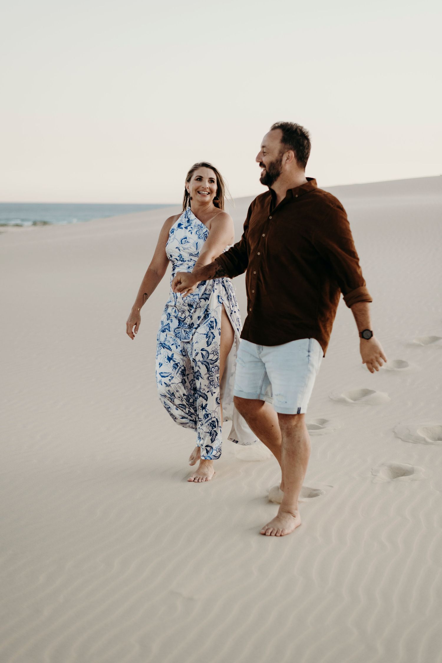 Couple running and playing together on scenic white sand dunes during a romantic photoshoot at sunset.
