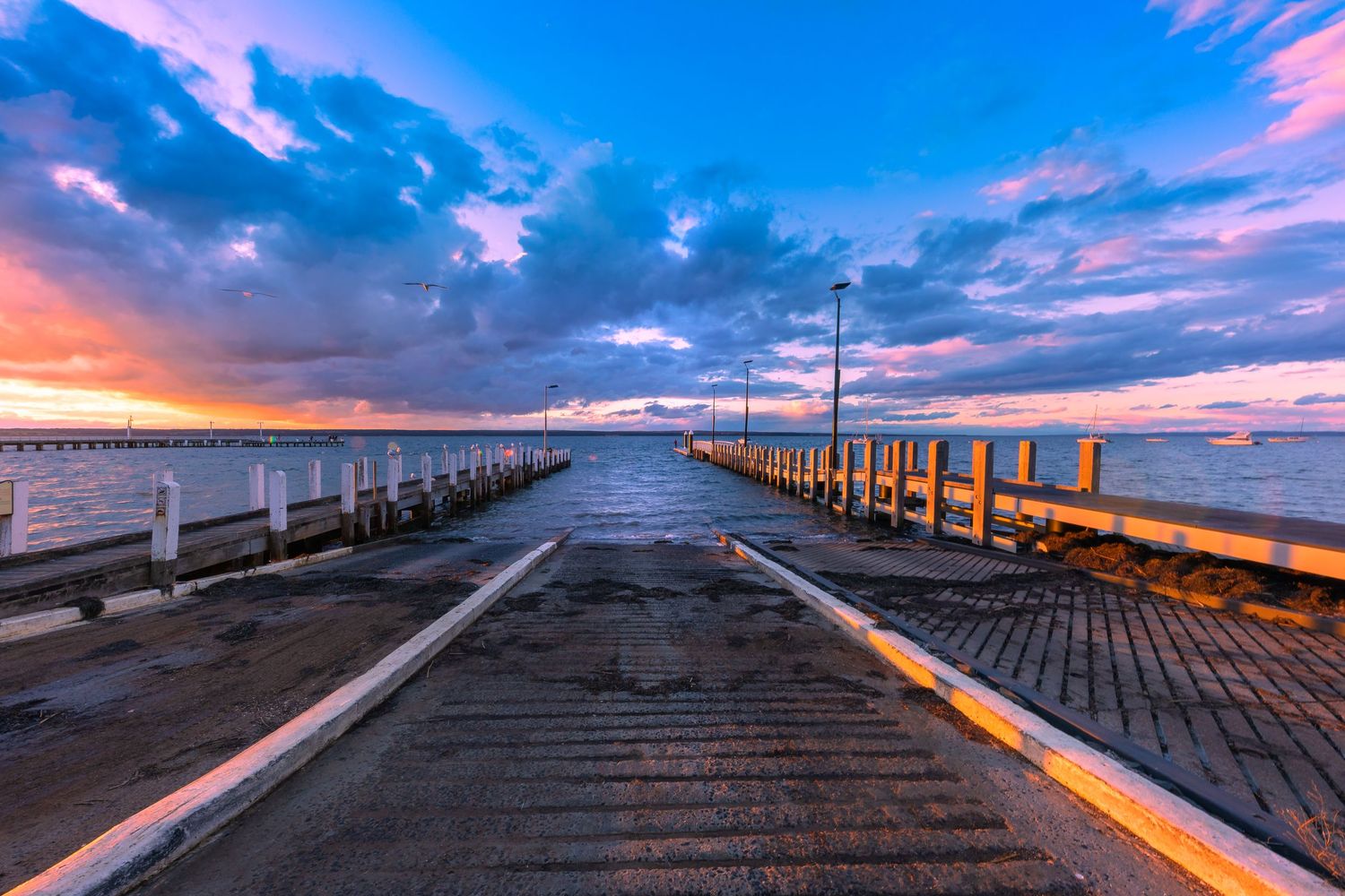 A wooden pier stretches into the ocean under a vibrant pink and blue sunset sky with dramatic clouds.