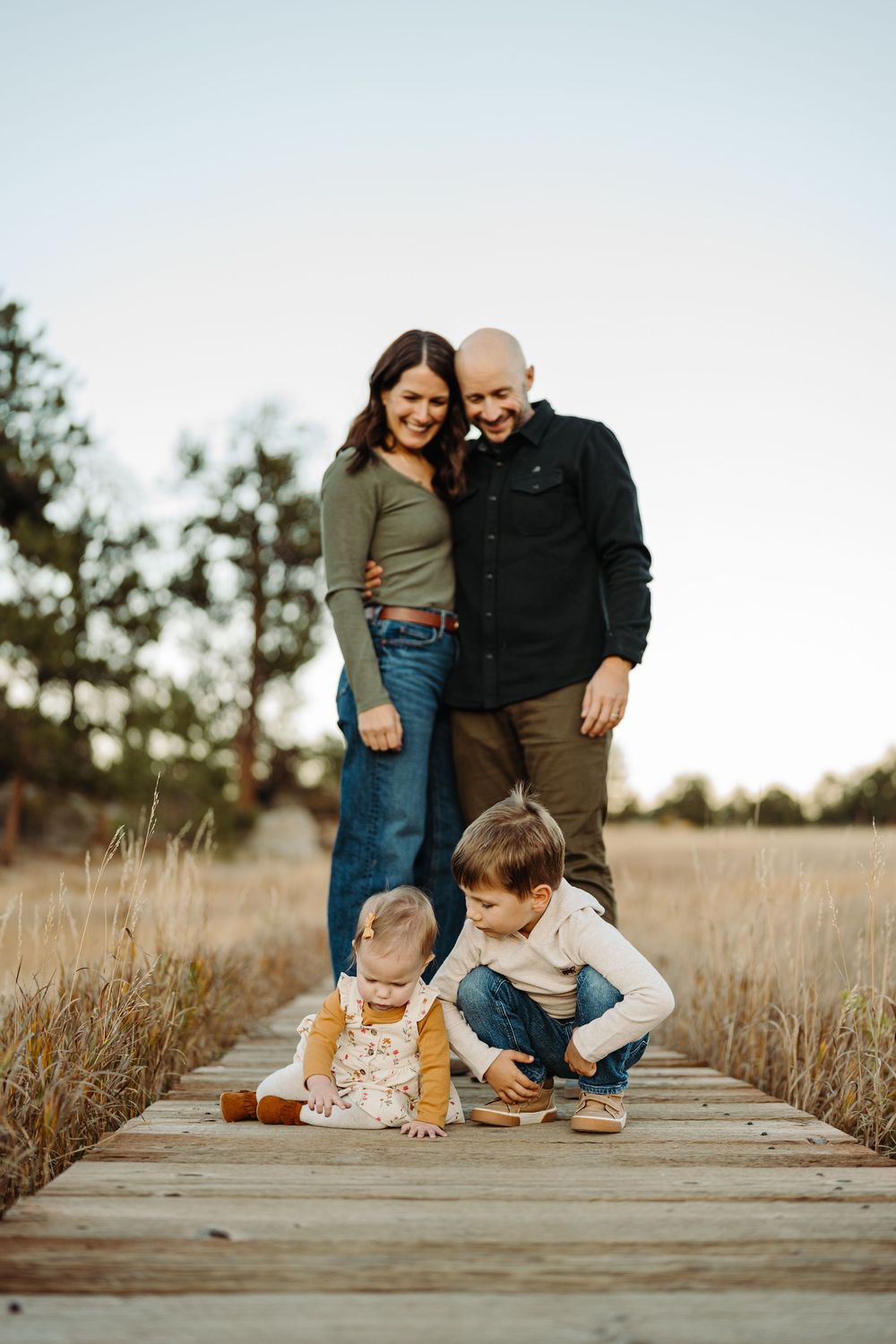 A family walks together on a wooden boardwalk through a field of tall grass at sunset.