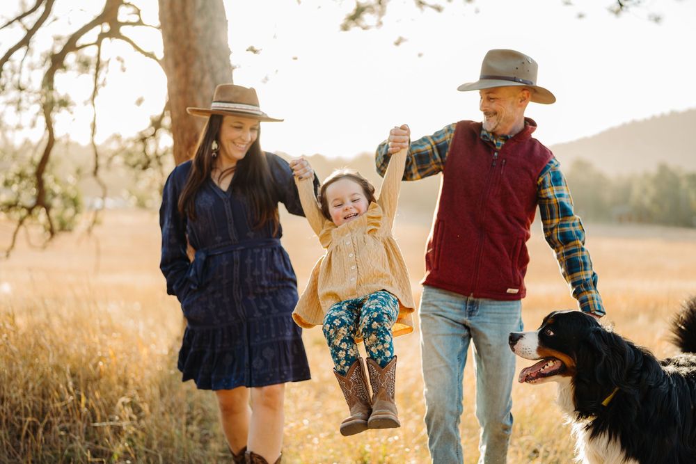 A family in warm autumn clothing explores a golden meadow at sunset with their black dog.