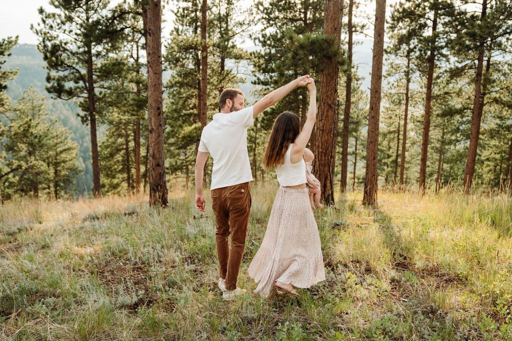 A couple holds hands while dancing together on a scenic hilltop surrounded by tall pine trees at sunset.