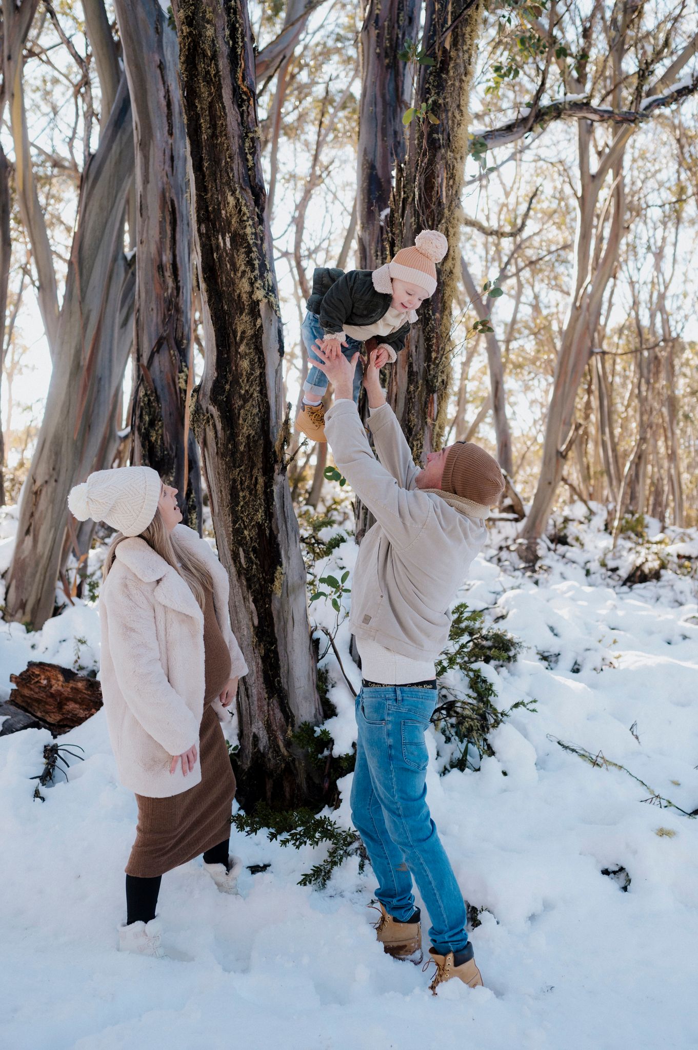 People in warm winter clothing play in a snowy forest with tall eucalyptus trees on a bright winter day.