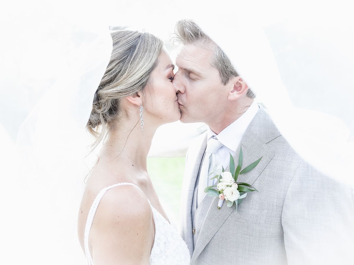A romantic wedding kiss captured in soft light with the groom wearing a light gray suit adorned with a white boutonniere.