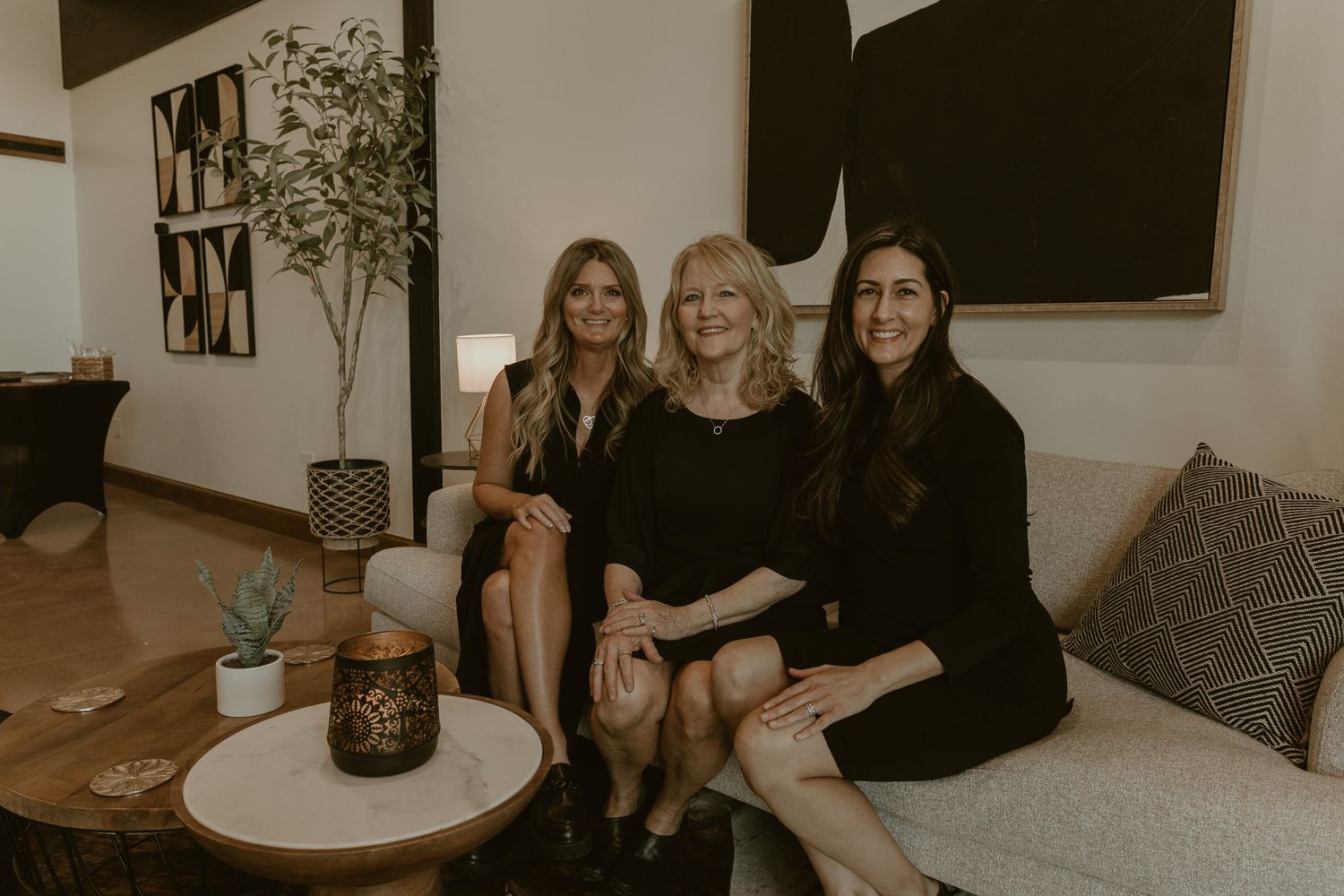 Three women in elegant black attire sit together on a couch in a modern room with minimalist decor.