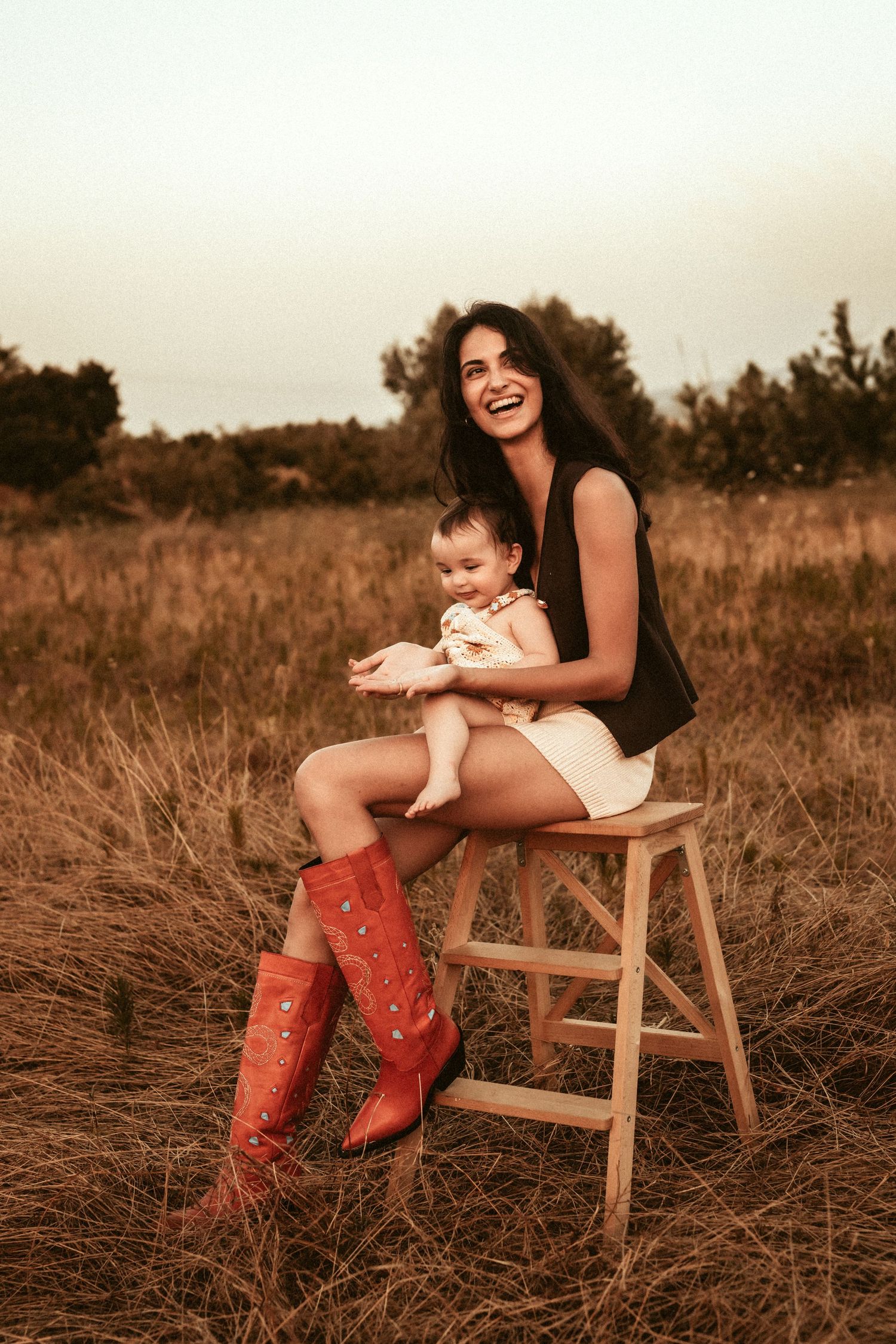 A woman in red cowboy boots sits on a wooden stool in a sunlit field holding an infant in Javea. Javea photographer